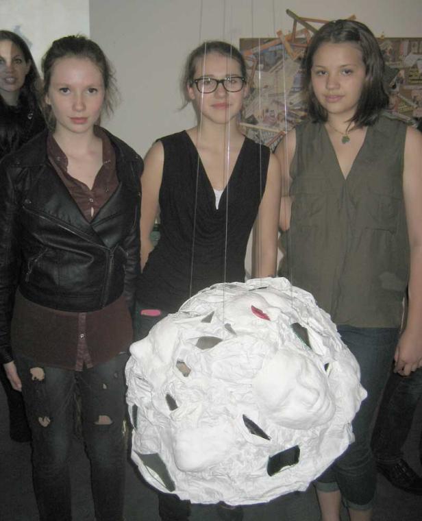 Drei junge Frauen stehen vor einer weißen, gesichtsförmigen Skulptur.