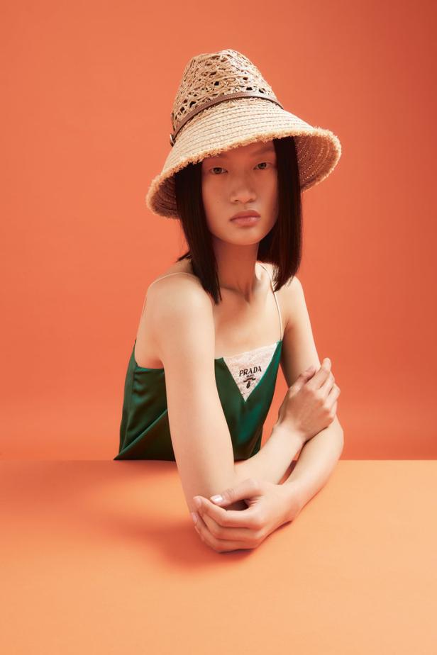Eine Frau mit Hut und einem grünen Prada-Top vor einem orangefarbenen Hintergrund.