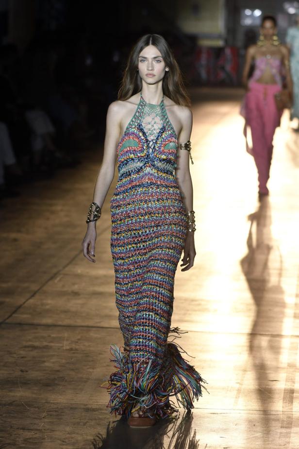 Ein Model präsentiert ein buntes, gehäkeltes Maxikleid mit Fransen am Saum auf einem Laufsteg.
