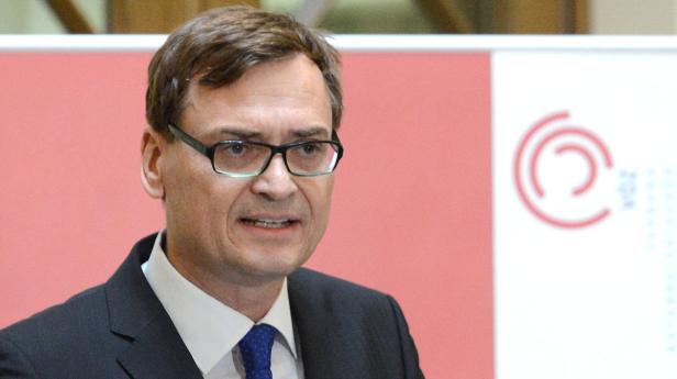 Ein Mann mit Brille und Anzug spricht vor einem roten Hintergrund.