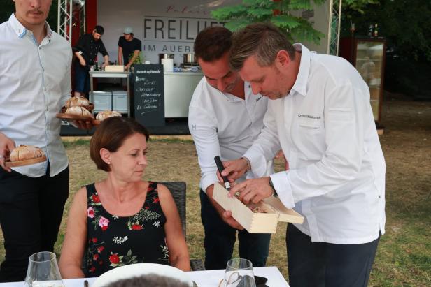 Ein Koch, möglicherweise Christof Widakovich, präsentiert einer Frau ein Gericht in einer Holzschatulle.