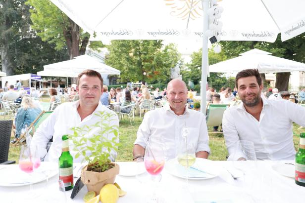 Drei Männer sitzen an einem Tisch im Freien unter Sonnenschirmen.