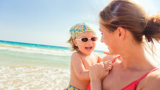 Eine Mutter hält ihr lachendes Kind mit Sonnenbrille und Kopftuch am Strand.