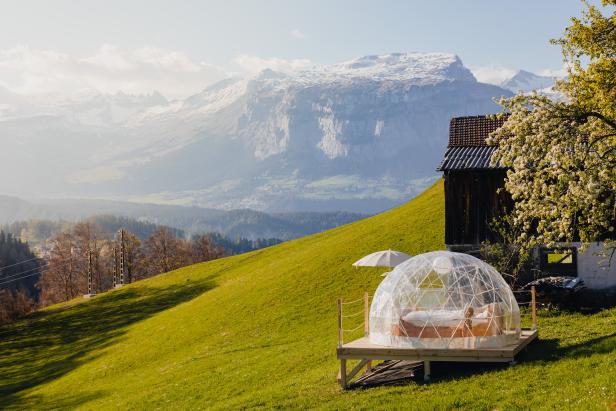 Ein durchsichtiges Iglu-Zelt steht auf einer grünen Wiese mit Blick auf die Berge.