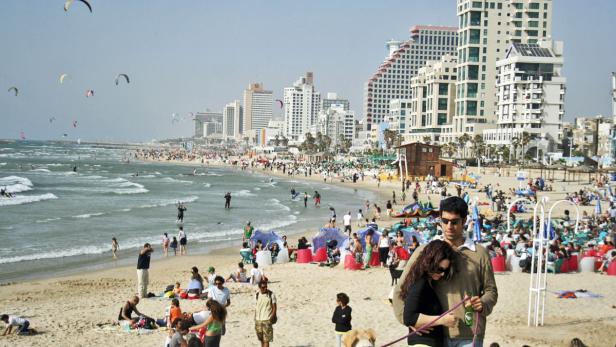 Ein belebter Strand in Tel Aviv mit vielen Menschen und Hochhäusern im Hintergrund.