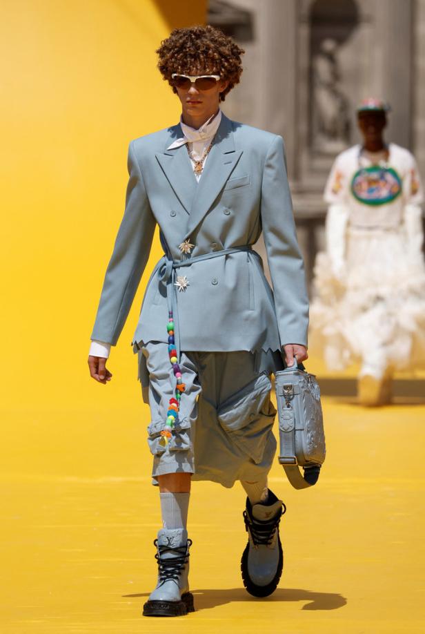 Ein Mann mit lockigem Haar trägt einen hellblauen Anzug und Stiefel auf einem gelben Laufsteg.