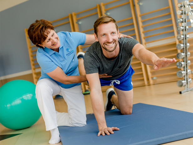 Ein Mann macht mit Unterstützung einer Physiotherapeutin Übungen auf einer Matte.