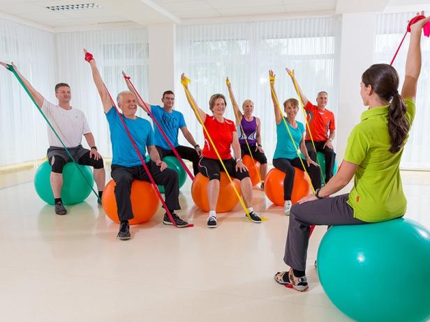 Eine Gruppe von Menschen trainiert mit Bändern auf Gymnastikbällen unter Anleitung einer Trainerin.