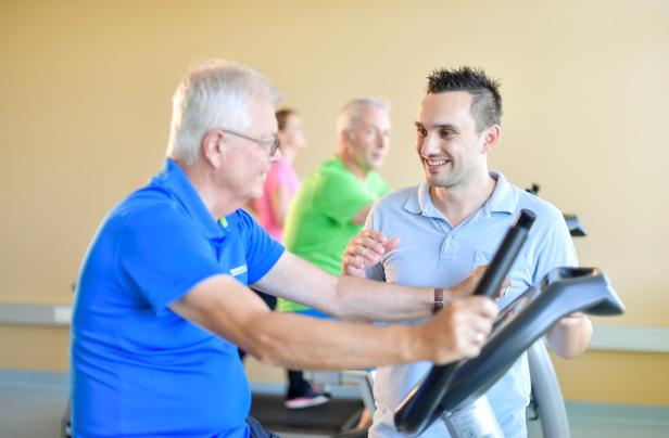 Ein älterer Mann trainiert mit Hilfe eines jüngeren Trainers an einem Fitnessgerät.