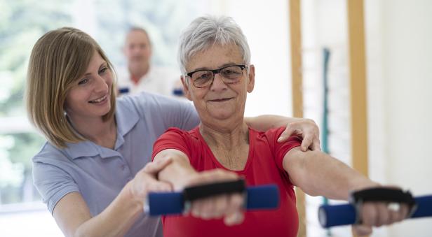 Eine ältere Frau macht mit Hilfe einer Physiotherapeutin Übungen mit kleinen Hanteln.