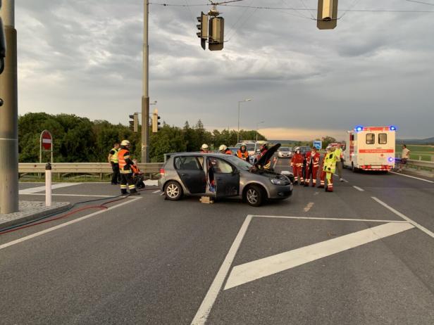 Ein Auto mit geöffneter Motorhaube steht auf einer Straße, umgeben von Rettungskräften und einem Krankenwagen.