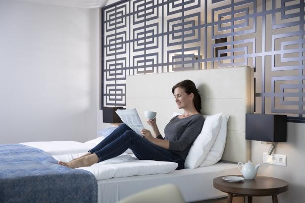 Eine Frau liest Zeitung und trinkt eine Tasse Tee im Bett eines Hotelzimmers.