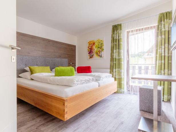 Ein helles Schlafzimmer mit Doppelbett, grünen Vorhängen und einem farbenfrohen Bild an der Wand.