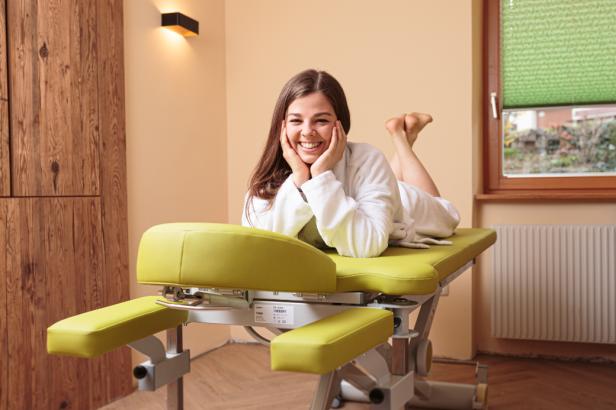 Eine lächelnde Frau liegt entspannt in einem Bademantel auf einer Massageliege.