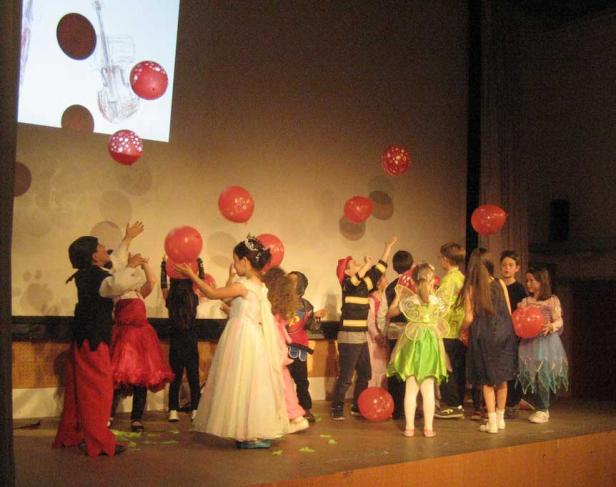 Eine Gruppe Kinder in Kostümen spielt auf einer Bühne mit roten Luftballons.