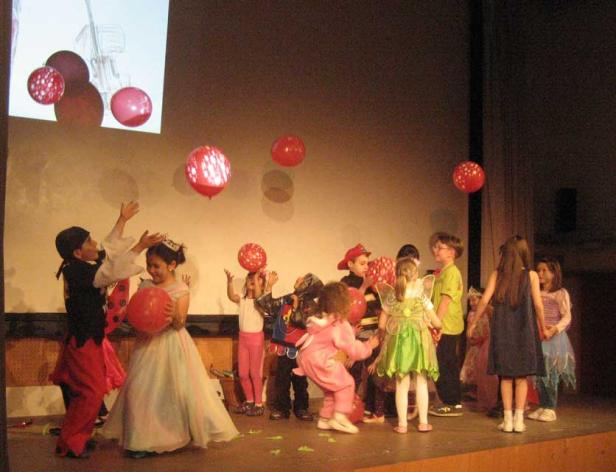 Eine Gruppe von Kindern in Kostümen steht auf einer Bühne mit roten Luftballons.