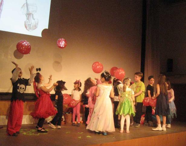 Eine Gruppe Kinder in Kostümen steht auf einer Bühne mit roten Luftballons.