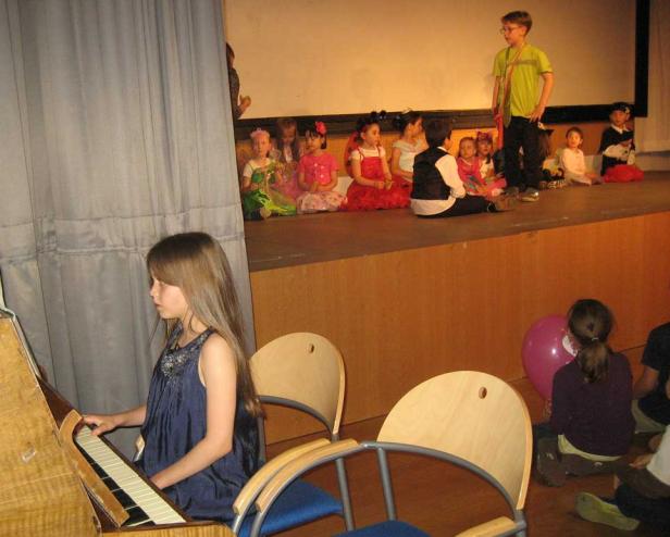 Ein Mädchen spielt Klavier, während andere Kinder auf einer Bühne sitzen.