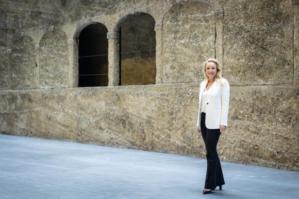 Eine Frau in Blazer und Hose steht vor einer Wand mit Torbögen.