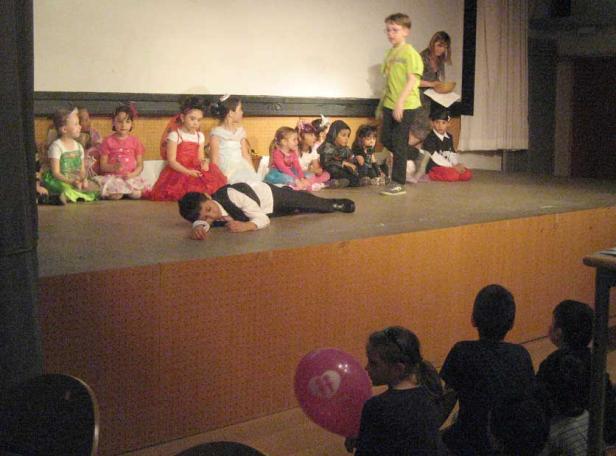 Eine Gruppe von Kindern sitzt auf einer Bühne, während ein Junge im Anzug auf dem Boden liegt.