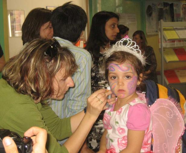 Ein Mädchen mit Krone und Gesichtsbemalung wird von einer Frau geschminkt.