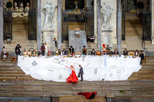 Eine Theaterszene mit einer langen Tafel und einer Frau im roten Kleid im Vordergrund.