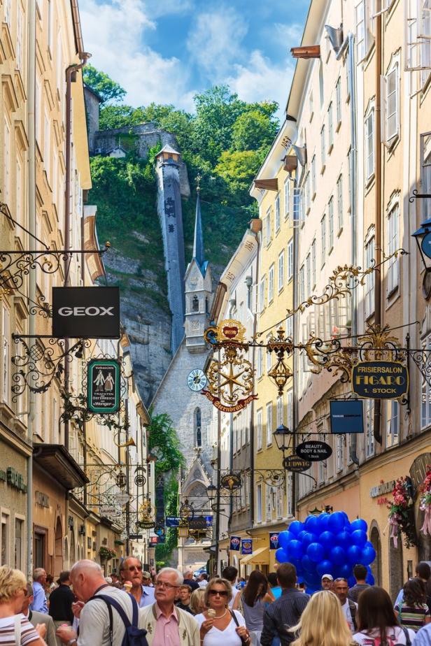 Blick durch eine belebte Gasse in Salzburg mit der Festung Hohensalzburg im Hintergrund.