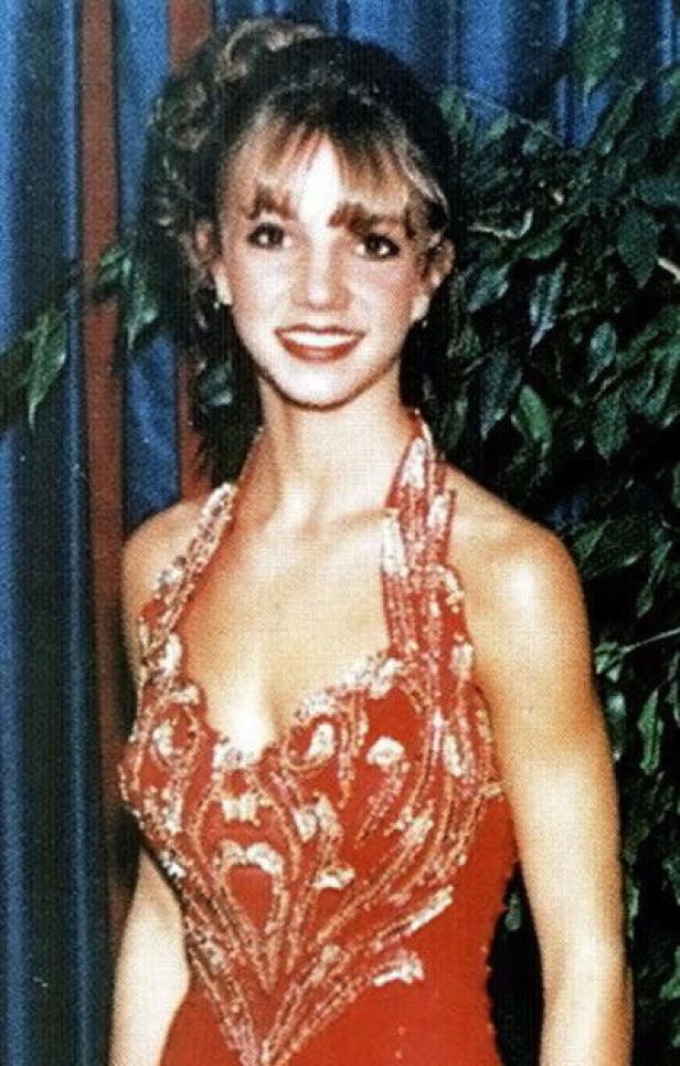 Britney Spears in einem roten, verzierten Kleid vor einem blauen Vorhang und einer Pflanze.