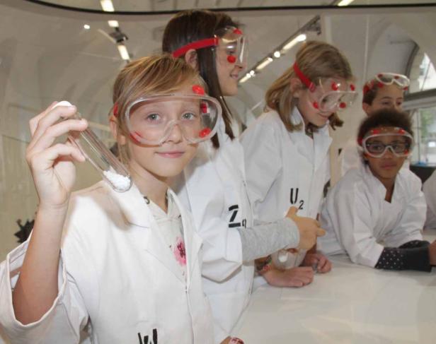 Eine Gruppe Kinder in Laborkitteln und Schutzbrillen experimentiert mit Chemikalien.
