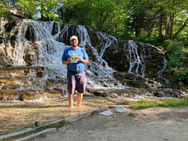 Ein Mann steht barfuß in einem Bach vor einem Wasserfall und hält ein Buch in der Hand.