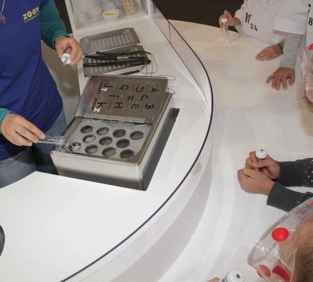 Eine Person füllt eine Flüssigkeit in eine Form mit Buchstaben, während Kinder zusehen.