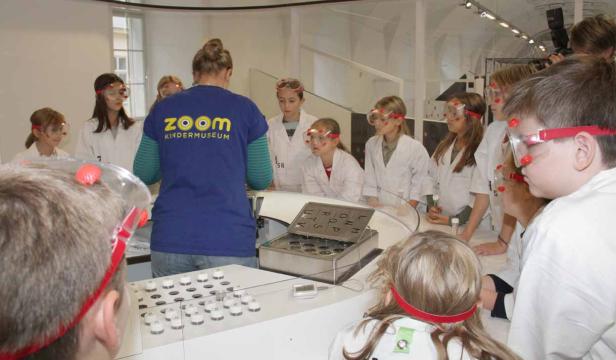 Eine Gruppe Kinder im Zoom Kindermuseum experimentiert mit Schutzbrillen und Laborkitteln.