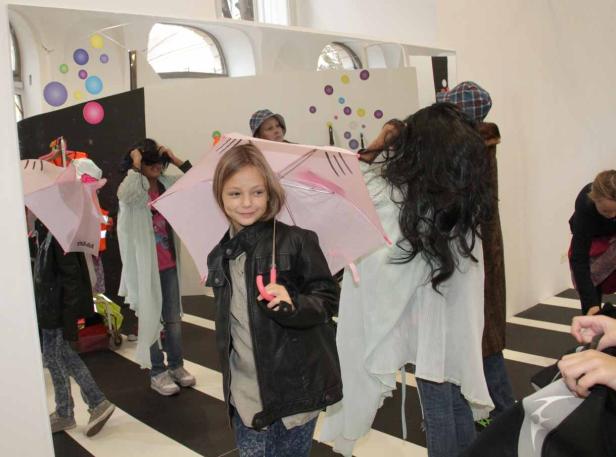 Ein junges Mädchen lächelt und hält einen rosa Regenschirm in einem Raum mit anderen Kindern.