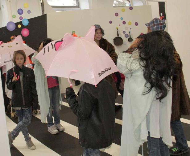 Mehrere Kinder stehen mit Hello Kitty-Regenschirmen in einem Raum mit gestreiftem Boden.