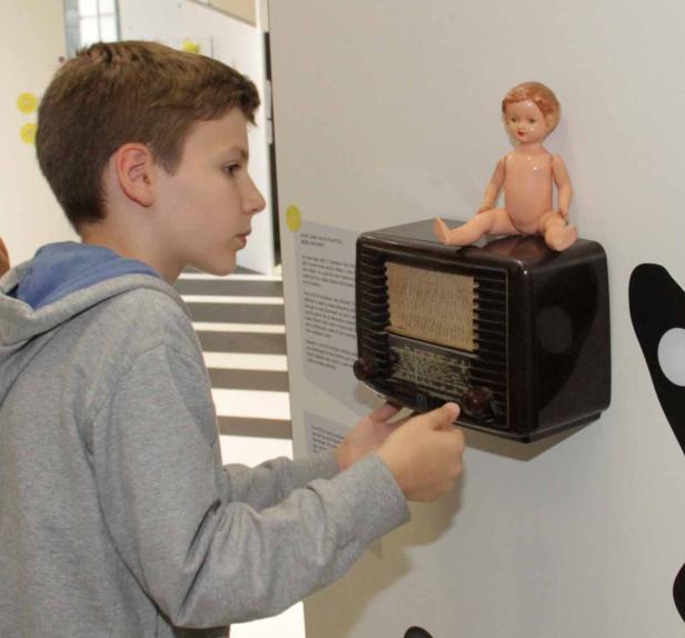 Ein Junge betrachtet ein altes Radio, auf dem eine Puppe sitzt.
