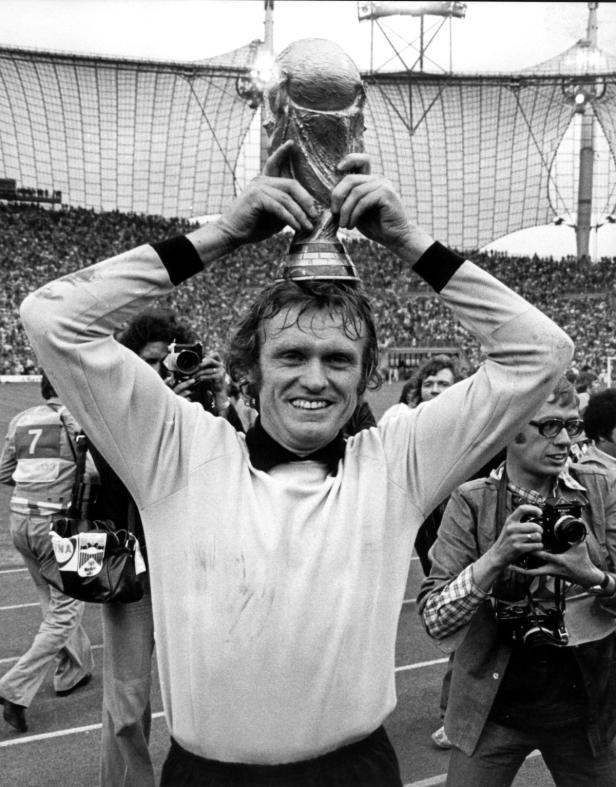 Franz Beckenbauer hält den Weltpokal auf seinem Kopf, umgeben von jubelnden Fans.