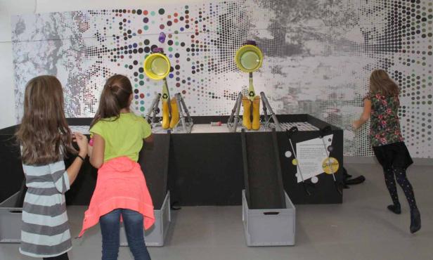 Drei Mädchen spielen mit einer interaktiven Installation mit gelben Gummistiefeln und bunten Punkten an der Wand.