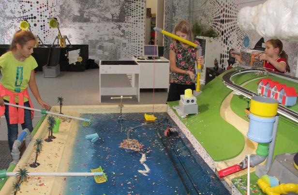 Zwei Mädchen spielen mit einem Modell, das eine Küstenlandschaft und ein Gewässer mit Plastikmüll darstellt.