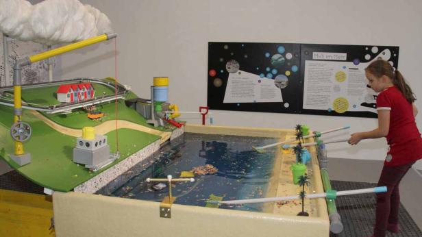 Ein Mädchen fischt mit einem Kescher Plastikmüll aus einem Wasserbecken, das Teil einer Installation zum Thema Umweltverschmutzung ist.