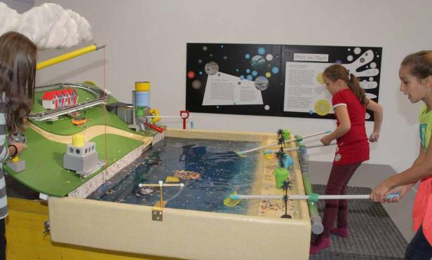 Zwei Mädchen fischen Plastikmüll aus einem Wasserbecken, neben einer Modelllandschaft.