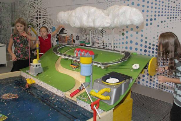 Drei Kinder spielen mit einem interaktiven Modell einer Landschaft mit Wasser, Bergen und einer Wolke.