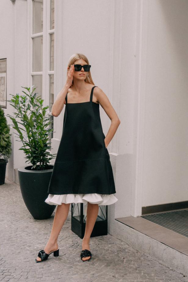 Eine blonde Frau mit Sonnenbrille trägt ein schwarzes Kleid mit Rüschen und schwarze Schuhe.