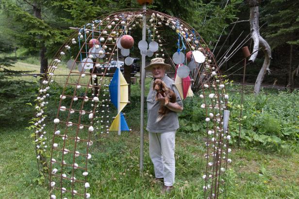 Ein älterer Mann mit Hut hält einen Dackel vor einer dekorierten Metallstruktur im Garten.