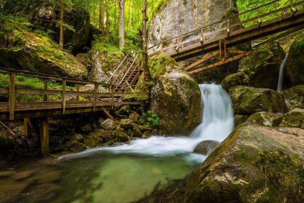 Ein Wasserfall fließt zwischen moosbewachsenen Felsen hindurch, überbrückt von Holzstegen.