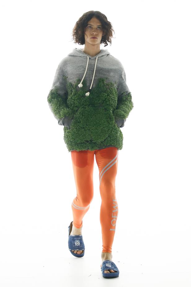 Ein Model auf dem Laufsteg in einem grauen Hoodie mit Pflanzenbewuchs und orangefarbenen Leggings von Loewe.