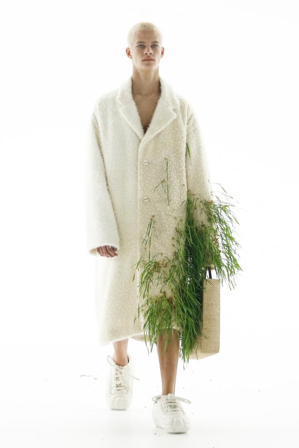 Ein Model mit blond gebleichten Haaren trägt einen weißen Mantel mit Gras und eine Tasche auf dem Laufsteg von Loewe.