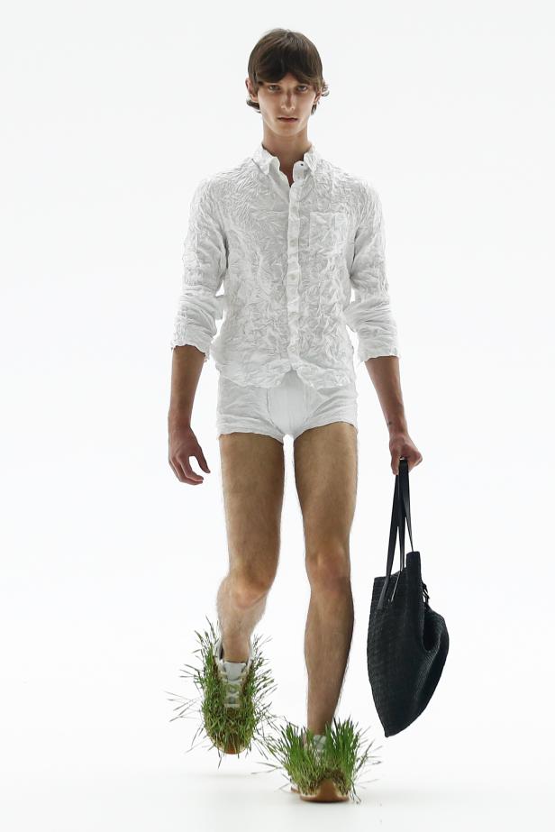 Ein Mann in weißem Hemd und Unterhose mit Gras an den Schuhen und einer Tasche.