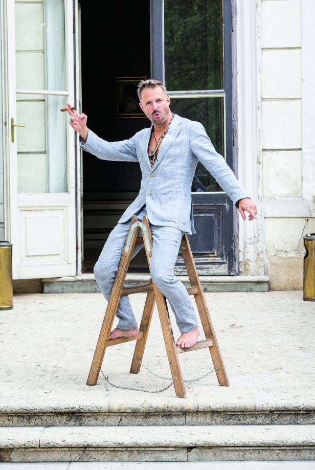Ein barfüßiger Mann im Anzug balanciert auf einer Leiter und raucht eine Zigarre.
