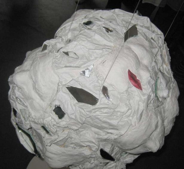 Eine weiße, zerknitterte Skulptur eines Kopfes mit Spiegelstücken und einem roten Lippenaufkleber.