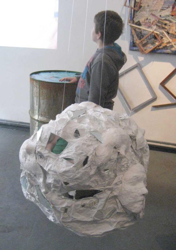Ein Junge betrachtet eine schwebende Skulptur mit Gesichtern und Spiegelstücken in einer Kunstausstellung.